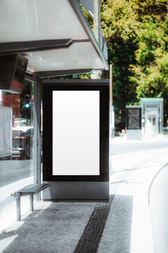 Blank vertical poster template inside a modern bus shelter with bench, bright sidewalk, road curve and green trees in soft urban background