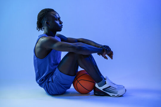 Young basketball player sitting with ball in blue studio light
