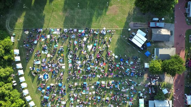 July Fourth Independence Day groups populate open park fields adjacent to Farmers Branch Historical Park stage. Expansive green spaces surround lively outdoor event area in Dallas Fort Worth, TX
