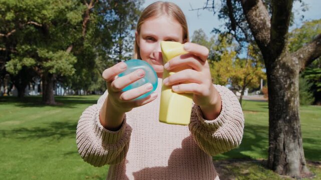 Young girl holds and squeezes two stress squishy toys, one blue and one yellow, while standing on grass in a park.