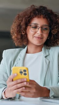 Smiling young indian or muslim arabian businesswoman in glasses using smartphone at desk in office. Mobile work communication. Latin curly young woman working on cellphone phone at workplace. Vertical