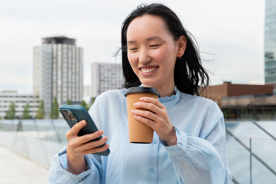Gen Z Asian woman with coffee using smartphone outdoors
