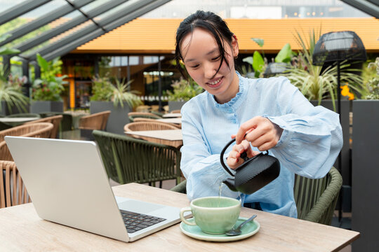 Young Gen Z Asian woman pouring tea at cafe with laptop