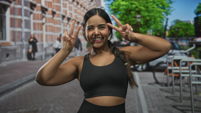 Young hispanic woman in black sportsbra flexing biceps and posing with double peace signs on a city street with brick building and bicycle racks; joy confidence.