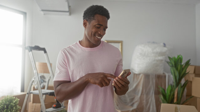 Man tapping smartphone with finger among packed boxes and ladder beside houseplant in building; moving day happiness.