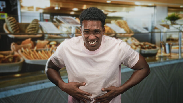 Man in pink tshirt clutching stomach with hands amid bakery pastries by bread display at building counter; pain nausea urgency.