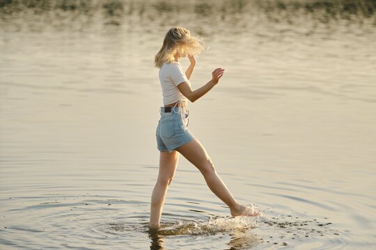 A woman walks through shallow water, splashing lightly as golden sunlight reflects off the surface. The calm setting creates a peaceful summer atmosphere during the evening hours