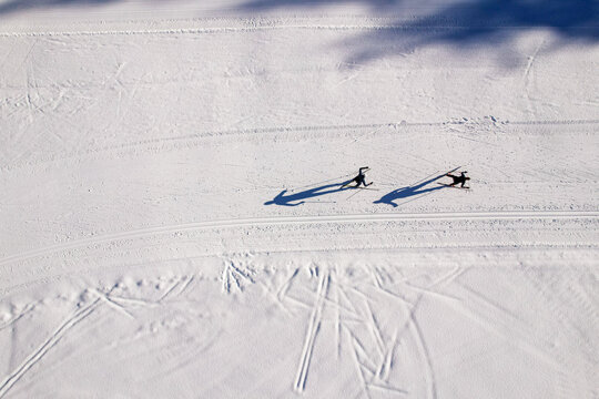 Aerial view of two cross-country skiers gliding along a groomed snow track with long shadows cast on the pristine white surface in Pragelato, Piedmont, Italy.