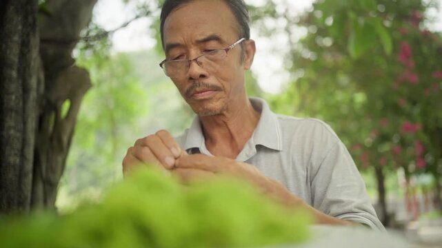 Elderly asian man with glasses carefully threading a needle with green cotton in tam coc, vietnam