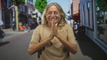 Fototapeta premium Senior woman clapping hands on a busy sunlit street, wearing a beige t shirt with bicycles and storefronts behind, smiling and laughing; joy gratitude.