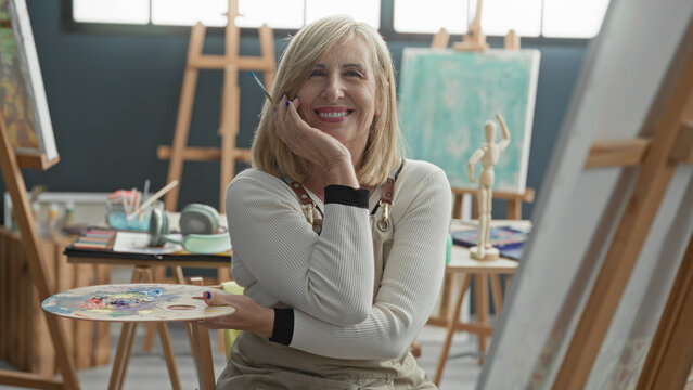 Middle aged blonde woman artist holds palette and brush, right hand resting on cheek while smiling in a bright art studio; creativity serenity.