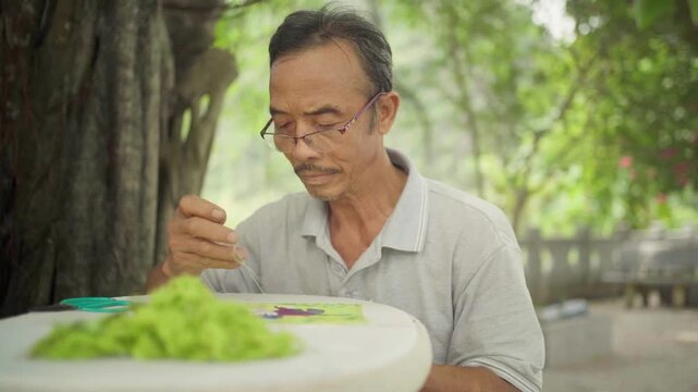 Elder asian man with glasses concentrating while hand sewing a colorful piece of traditional fabric