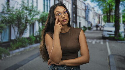 Fototapeta premium Woman touching eyeglasses with finger on cheek in street while leaning with arms crossed; pensive curiosity.