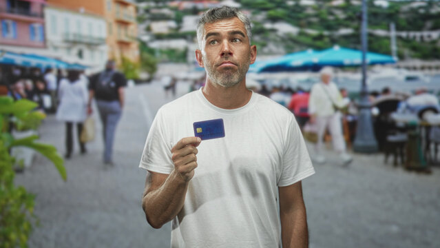 Man holding creditcard in right hand on a busy street with cafes and waterfront backdrop, grey middleaged male faces camera; financial concern.