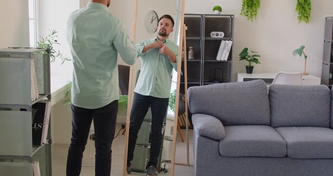 At home, a man in a bright livingroom adjusting his shirt and checking his outfit in a full-length mirror, focusing on reflection, grooming, and fashion. Man preparing for work in the morning