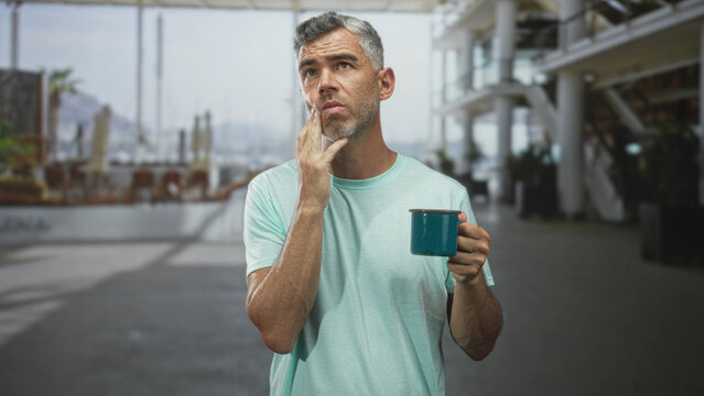 Man holding teal mug presses cheek with wince and closed eyes for toothache inside a bright building concourse; discomfort morning routine.