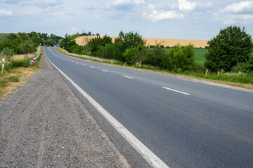 Fototapeta premium a clear, paved two-lane asphalt highway stretching into the distance through a rural landscape with green trees and golden fields under a partly cloudy sky.
