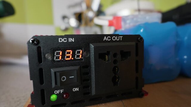 A close-up of a red power inverter displaying voltage on its digital LED screen. Concept of off-grid living, alternative green energy, and portable battery power supply.