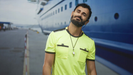 Fototapeta premium Man in middle age with beard wearing a fluorescent referee shirt, whistle on lanyard and headset, smiling with eyes closed and head tilted at cruise terminal building pier; contentment.