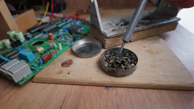A technician dips a hot soldering iron into a small container of dark rosin flux on a wooden table. A green printed circuit board lies nearby under soft indoor light.