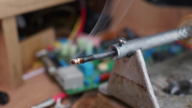 A hot soldering iron tip emits smoke as melted rosin and solder burn on a workbench stand. Concept of electronics repair, hardware engineering, DIY hobby, and manual labor.