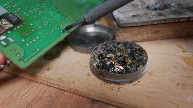 Hot soldering iron tip dips into dark rosin beside a green printed circuit board on a wooden desk. Concept of electronics repair, hardware maintenance, and DIY engineering.