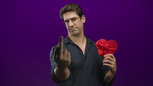 Man in dark shirt holds a red satin heart gift with bow while showing middle finger toward camera in purple studio; defiance rejection.