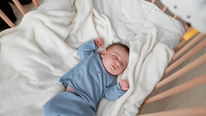 Baby sleeping with clenched fists and one hand near face on a soft blanket in a wooden crib inside a building  peaceful rest. © Krakenimages.com
