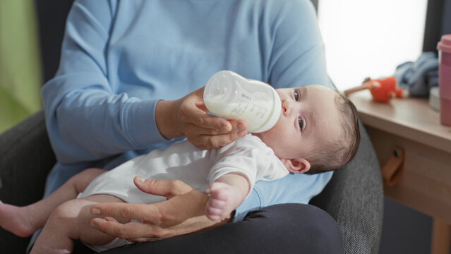 Newborn boy fed from a bottle while caregiver holds him and offers milk at a chair in a building; comfort bonding.