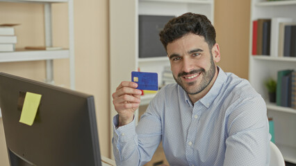 Young hispanic man holds a blue creditcard with his hand near a computer monitor smiling inside a building office with bookshelves and a sticky note on the screen  satisfaction. © Krakenimages.com