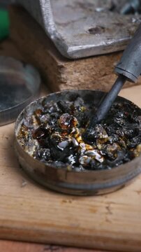 A hot soldering iron tip melts dark rosin flux inside a metal tin container. Wisps of smoke rise as the heated tool liquefies the amber colophony resin on a wooden desk.