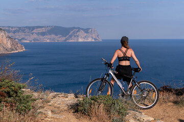 Fototapeta premium Bicycle travel woman standing on a cliff overlooking the ocean with a scenic mountain landscape view in the distance