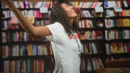 Teenage woman lifeguard with curly hair wearing a whistle, arms out smiling by crowded bookshelves in building  joy freedom. © Krakenimages.com