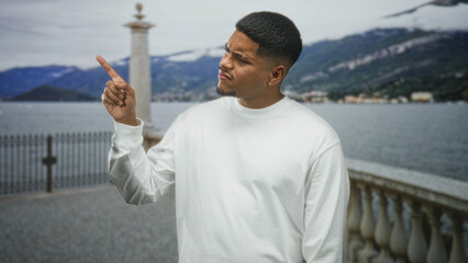 Fototapeta premium Hispanic man with beard in white sweatshirt points index finger upward on a lakeside street by a stone balustrade and iron railing with mountains across the lake; confidence.