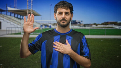 Naklejka premium Man in blue football shirt raises right hand palm outward and places left hand on chest in building near a soccer pitch and spectator stands; pride.