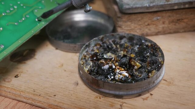 A hot soldering iron tip is dipped into a tin container filled with dark rosin flux, releasing smoke. Melted amber colophony bubbles around the heated metal tool.