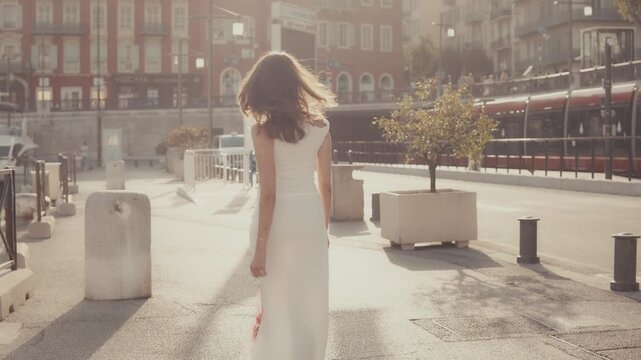 Back view of a young beautiful woman in a long white floral dress walking away along a sunny city street in europe, with her hair blowing in the wind during a golden hour sunset, slow motion