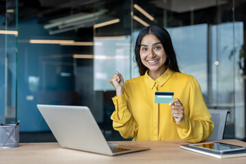 Fototapeta premium Happy young Indian woman sitting at office desk, holding credit financial card in hand and showing success gesture