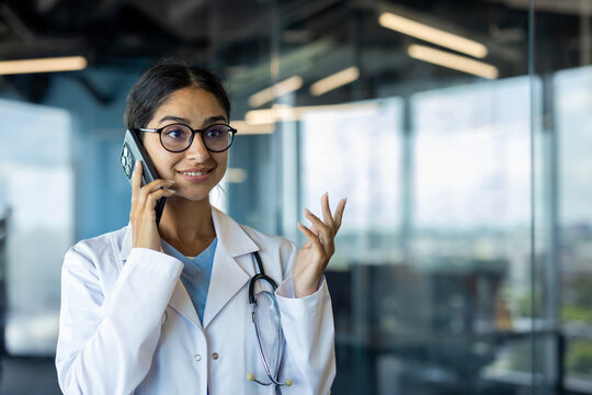 Smiling young Indian doctor standing in clinic office and talking on phone