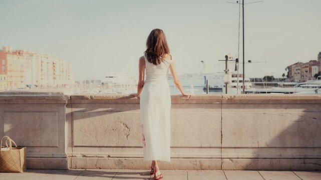 Stylish brunette woman wearing a long white dress and leaning on a stone balustrade while admiring the beautiful view of a mediterranean harbor filled with yachts on a sunny summer day