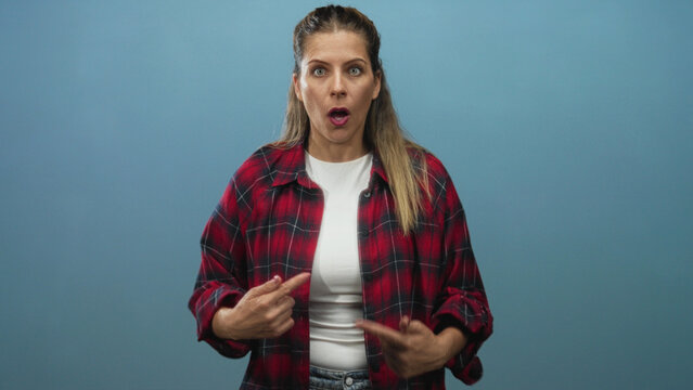 Woman in red plaid shirt pointing at herself with both thumbs and wide open mouth in studio; surprise disbelief.
