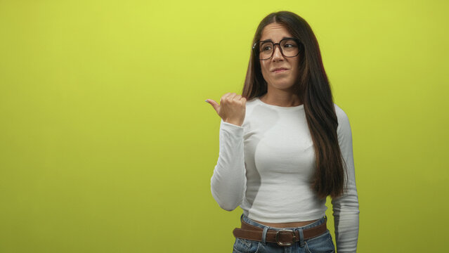 Woman points thumb over shoulder in bright lime studio, long hair and glasses, white long sleeve top and jeans belt visible; skepticism.