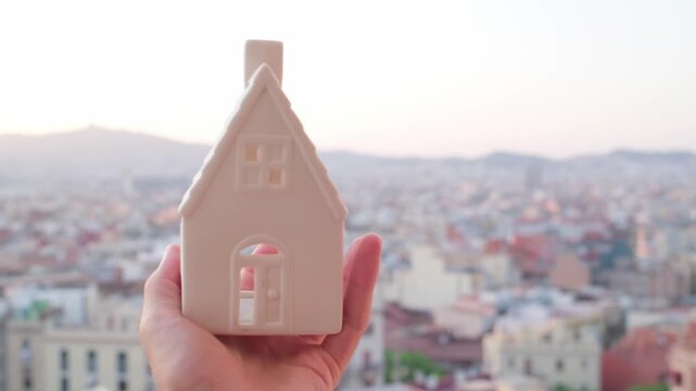 point of view of a hand holding a small house model against a blurred urban cityscape, symbolizing concept like real estate, home ownership, property investment, and urban housing planning