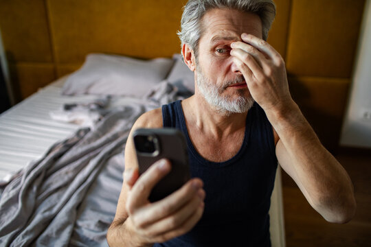 Middle-aged man rubbing eyes while checking smartphone in bed at home