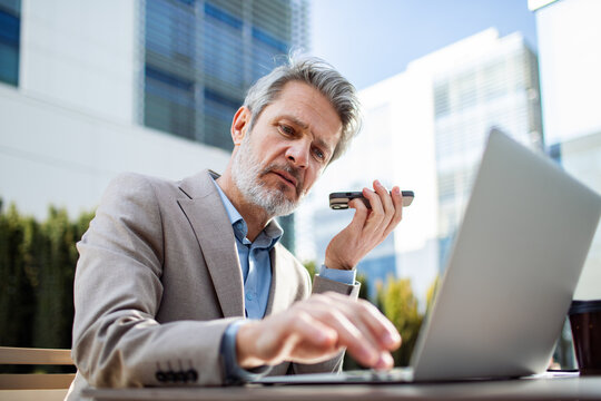 Senior businessman using voice assistant on smartphone while working on laptop outdoors