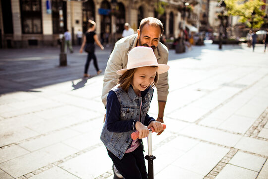 Father pushing daughter on scooter in sunny city street