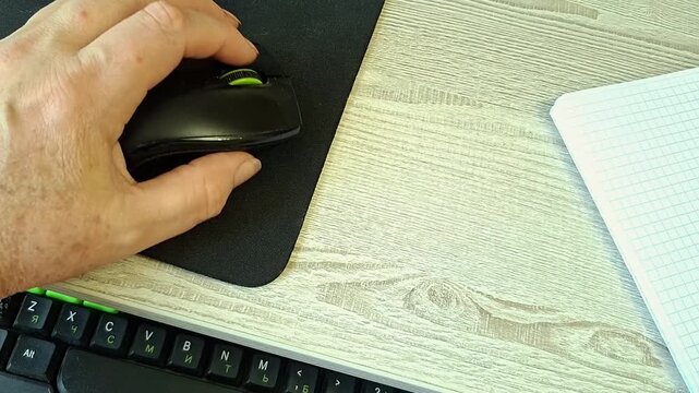 Senior person's hand using computer mouse at ergonomic desk with notebook
