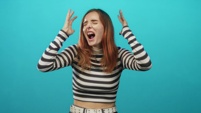 Young redhead woman in black and white striped top clenches fists, grimaces and squeezes eyes shut while centered against a turquoise studio backdrop; frustration.