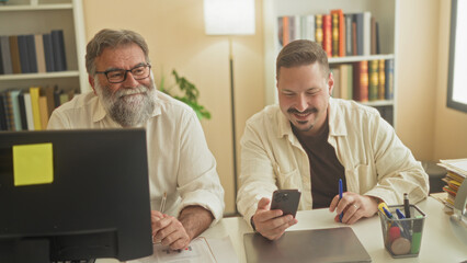 Two men at a desk view a smartphone and laptop, one man taps the phone screen while the other watches closely in a studio office with bookshelves and a lamp  focused collaboration. © Krakenimages.com