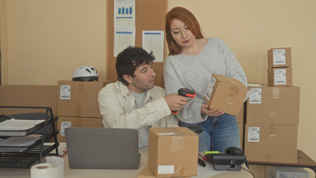 Man scans parcel with handheld scanner while seated at laptop in packing building; small business teamwork happy.